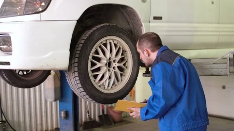 Car mechanic is checking breaks and writing in notepad on lifted white car Stock Footage 74889561