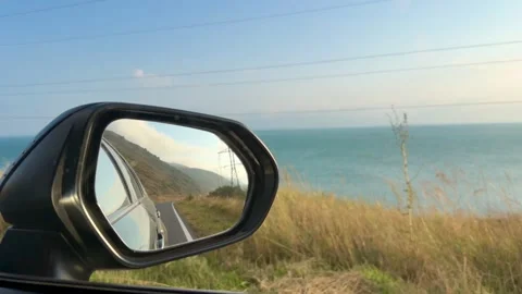 Car side mirror reflecting coastal scenery with ocean, road, and power lines Stock Footage 344722926