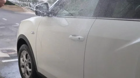 A car wash worker washes dirt off a white car using a high pressure washer with Stock Footage 256077163