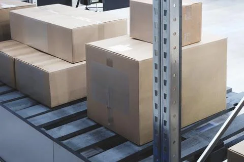 Cardboard boxes stack on conveyor belt in warehouse. Metal rack holds boxes s Stockfoto's