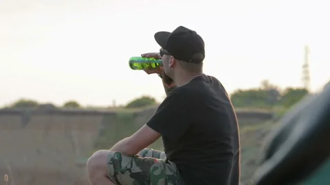 A carefree, young, friendly dude sits on a rock above the sea and drinks beer. Stockbeeldmateriaal 93485809