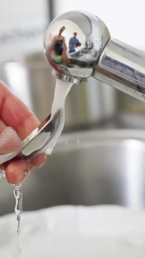 Carefully cleaning a spoon under running water in the kitchen for hygiene and Stock Footage 310466213