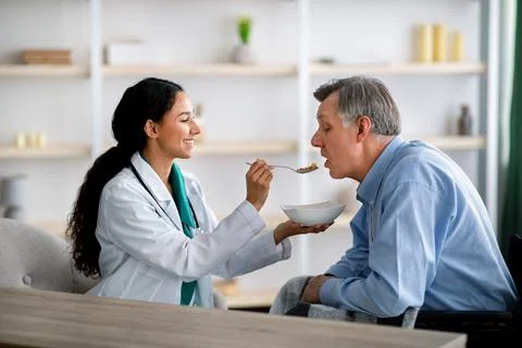 Caregiver feeding elderly man in wheelchair at home. Medical assistance for Stock Photos