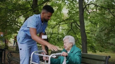 Caregiver man helping senior woman with walker to stand up from bench outdoors Stock Footage