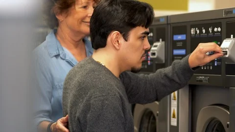 Carer helping a young man with a disability to do laundry at a launderette. Vídeos de archivo 88374996