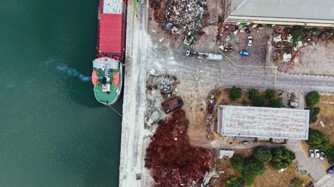 Cargo ship is docked near a site with scrap materials and a storage building by Stock Footage 341571445