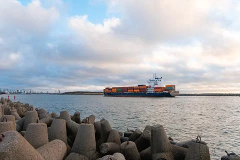 Cargo ship navigating through a rocky harbor entrance under a cloudy sky, s.. Stock-Fotos