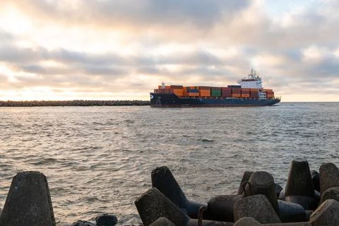 Cargo ship navigating through a rocky harbor entrance under a cloudy sky, s.. Stock Photos