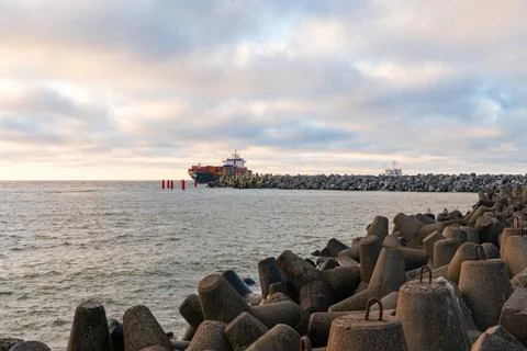 Cargo ship navigating through a rocky harbor entrance under a cloudy sky, s.. Stock Photos