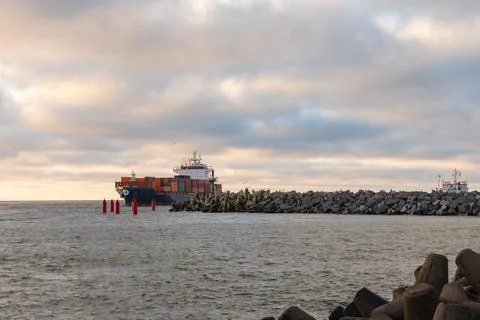 Cargo ship navigating through a rocky harbor entrance under a cloudy sky, s.. Stock Photos