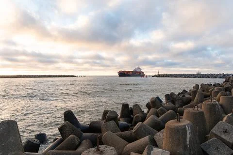 Cargo ship navigating through a rocky harbor entrance under a cloudy sky, s.. Stock Photos