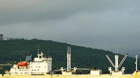 Cargo ship navigating under a cloudy sky near a green hillside Stock Photos