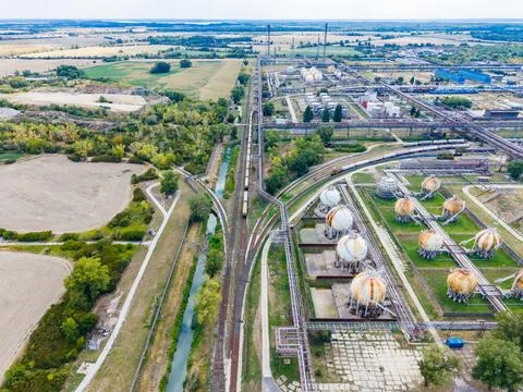 A cargo train moves along tracks in an industrial area with storage tanks and Stock Photos