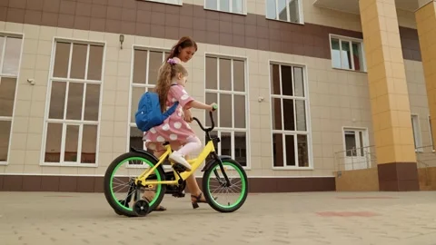 Caring young mother learning help little daughter riding bike at school building Stock Footage 280365198