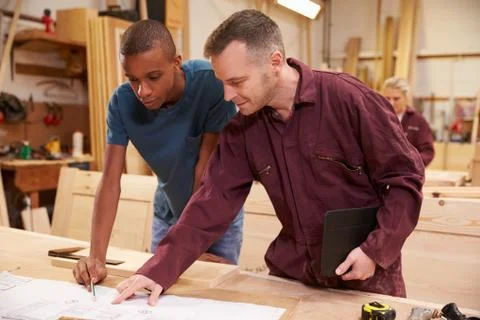 Carpenter With Apprentice Looking At Plans In Workshop Foto stock