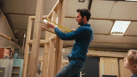 Carpenter With Female Apprentice In Workshop Using Retractable Ruler To Measure Stock Footage 260583600