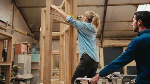 Carpenter With Female Apprentice In Workshop Using Retractable Ruler To Measure Video stock 260583743