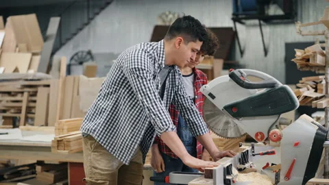 Carpenter teaching pretty girl apprentice to use electric saw in workshop Stock Footage 113745611