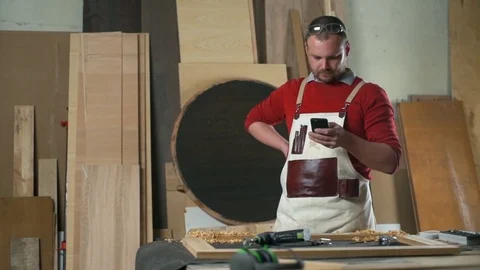 Carpenter wearing a woodworking apron using his smartphone in a workshop Stock Footage 74508344