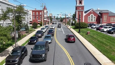Cars queue along narrow tree lined street beside red brick church with bell Stock Footage 343011122