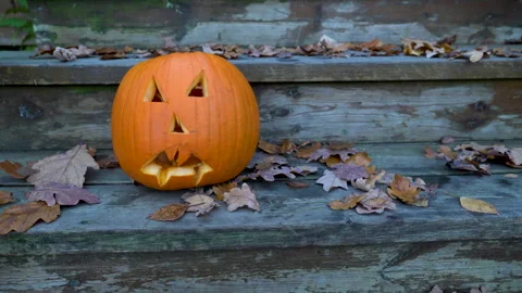 Carved pumpkin jack-o'-lantern on rustic wooden steps with falling leaves Stock Footage 289048046