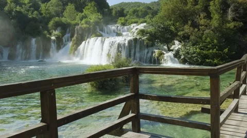 Cascading waterfall surrounded by lush trees and a rustic wooden railing. Видео 259074983