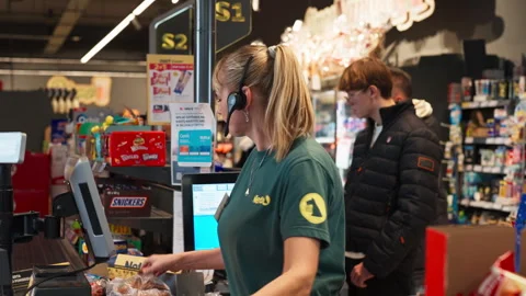 A Cashier Wearing a Headset Scans Items at the Checkout While Customers Wait in Stock Footage 325946590