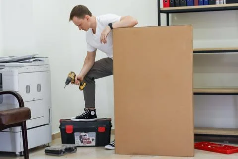Casual man on toolbox using drill on tall cardboard box indoors Stock Photos