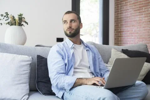 Casual man using laptop on sofa at home looking thoughtful while working re.. 写真素材