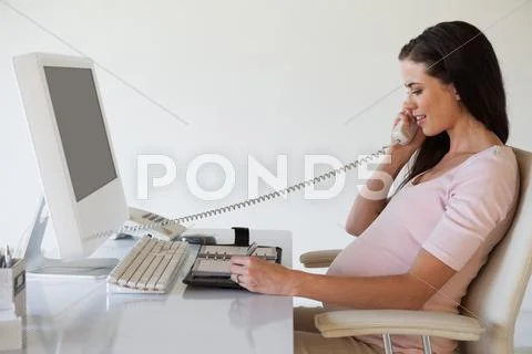 Casual pregnant businesswoman talking on phone at her desk Stock-Fotos