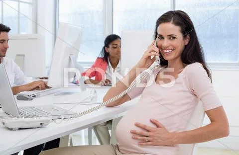 Casual pregnant businesswoman talking on phone at her desk Stock Photos
