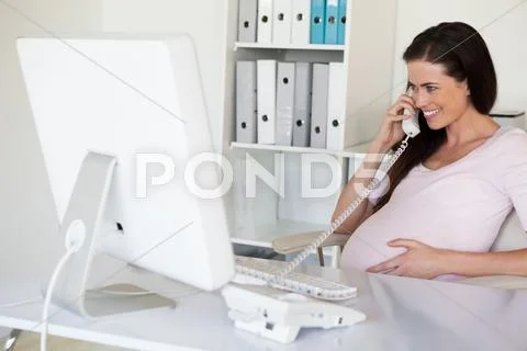 Casual pregnant businesswoman touching her bump at her desk talking on phone Stock Photos