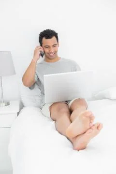 Casual smiling man using cellphone and laptop in bed Stock Photos