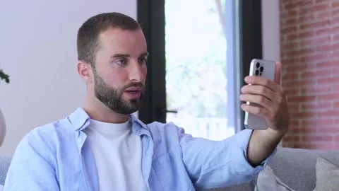 Casual young bearded man sitting on a sofa in his living room, having Stock Footage 345123889