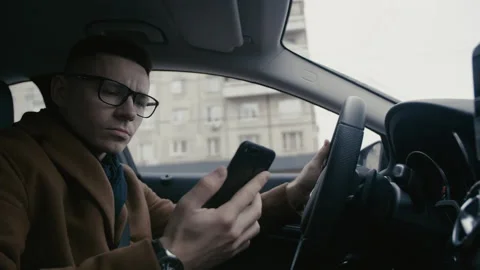 Casual young man in coat using smart phone while driving car on road. He is Stock Footage 156597782