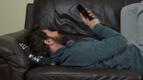 Casual Young Man Lying on Leather Sofa and Using Smartphone Video stock 76522662