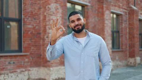 Casually dressed indian man winking and demonstrating okay gestures outdoors Stock Footage 250386466