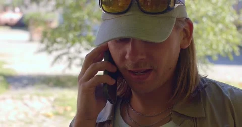A casually dressed man in a cap talks animatedly on his phone outdoors Stock Footage 325671124