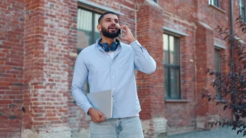 Casually dressed man with earbuds on neck holding laptop and talking on mobile Video stock 250388795