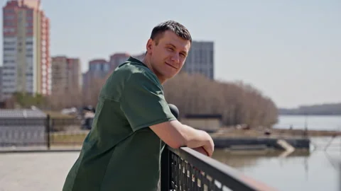 Casually dressed man leaning on riverfront railing with urban residential Stock Footage 274204909