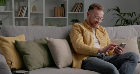 Casually dressed man sitting on cozy sofa while using smartphone. Video stock 249465796