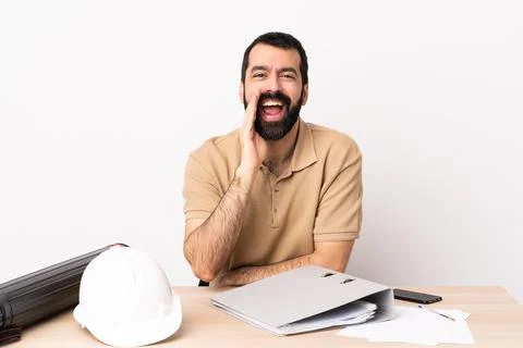 Caucasian architect man with beard in a table shouting with mouth wide open Photos