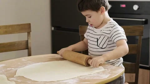 Caucasian boy rolls out dough with rolling pin on table in kitchen helping Stock Footage 192531358