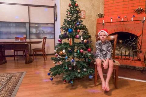 Caucasian boy sitting on chair wearing Santa hat near Christmas tree Stock Photos