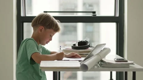 Caucasian boy sitting at table with workbook and looking at tablet screen while Video stock 194989754