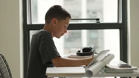 Caucasian boy sitting at table with workbook and looking at tablet screen while Stock Footage 194989936