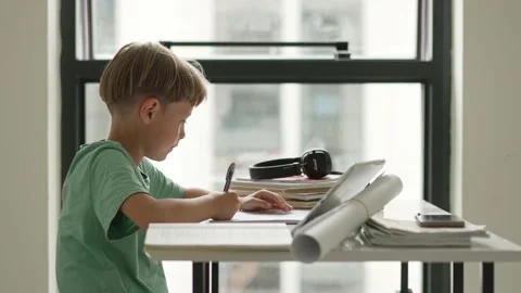 Caucasian boy sitting at table with workbook and looking at tablet screen while Stock-Footage 195098840