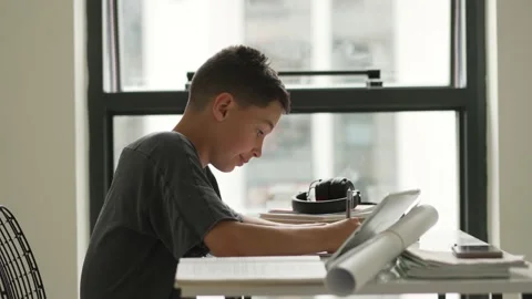 Caucasian boy sitting at table with workbook and looking at tablet screen while Stockbeeldmateriaal 195122582