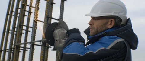 Caucasian builder in blue jacket and white helmet uses angle grinder, sparks Stock Footage 119496668