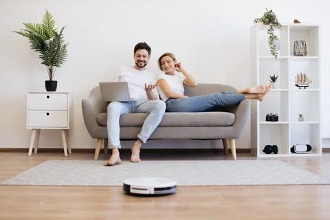Caucasian couple using laptop while robot cleaning floor Stock Photos